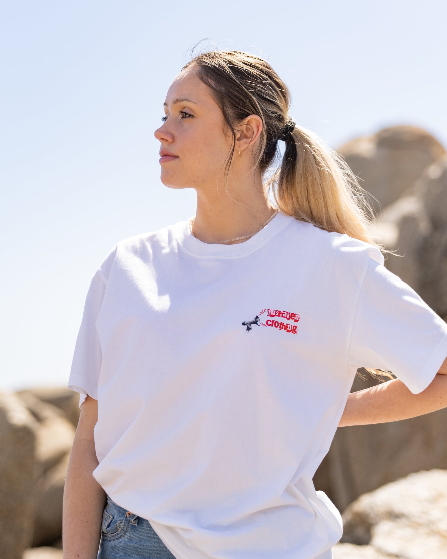 Woman wearing a white t-shirt with a logo, standing outdoors with rocks and clear sky in the background